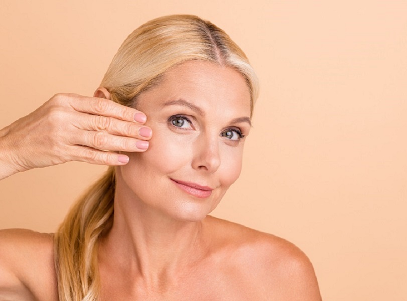 Closeup photo of natural beauty aged lady nude shoulders touch fingers cheek apply anti age uplift cream isolated beige color background | Kay Dermatology in Burbank, CA