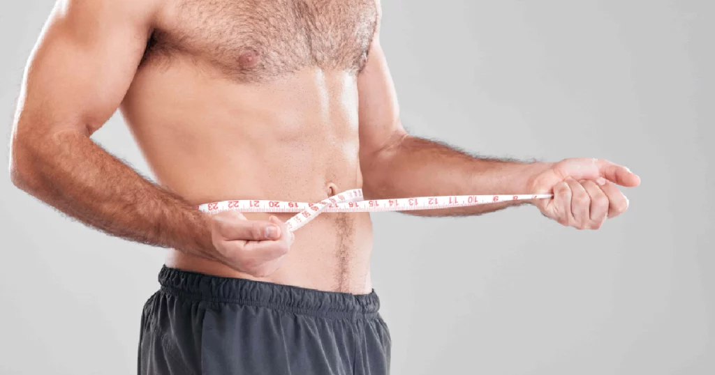 What to Expect Before, During, and After Male Liposuction in Burbank, CA 3 Shirtless man measuring his waist with a tape measure, showcasing a toned abdomen—representing Male Liposuction results and body contouring in Burbank, CA.